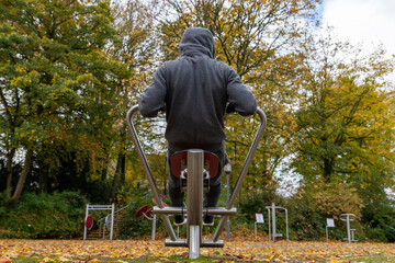 Ein Mann im Hoodie mit einer Kapuze auf dem Kopf trainiert an der Rudermaschine drau&szlig;en im Park im Herbst, R&uuml;ckenansicht..