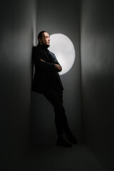 Fototapeta premium Calm sad handsome stylish man with long hair and beard in suit leans diagonally against the wall and looks thoughtfully down. Fashion style posing. Dark colors vertical composition with spotlight