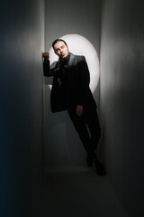 handsome stylish man with long hair and beard in suit leans diagonally against the wall and looks thoughtfully into the camera. Fashion style posing. Dark colors vertical composition with spotlight