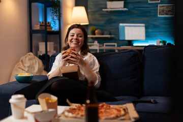Woman laughing watching comedy television series while eating tasty takeout burger relaxing on...