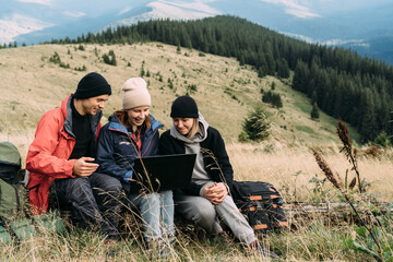Naklejka na ściany i meble a group of tourists three people a man and two women while relaxing in the mountains sit on a fallen tree and look into a laptop. Technology concept and coverage of 6w wireless networks.
