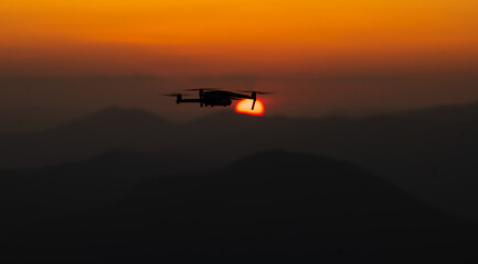 Fototapeta na wymiar Drone in flight. Silhouette of an UAV quadcopter flying in front of the sun during sunset on top of the mountains. Drones industry.
