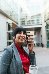 latina businesswoman talking on the phone and holding a recyclable cup of coffee on the street.