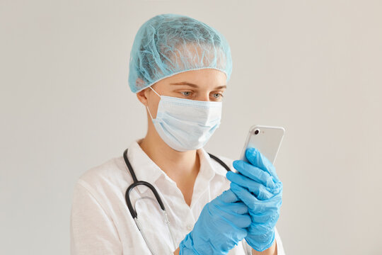 Image of attractive young adult woman doctor wearing medical cap, surgical mask, gown and rubber gloves, being at work in hospital, using mobile phone, reading message or information in the internet.