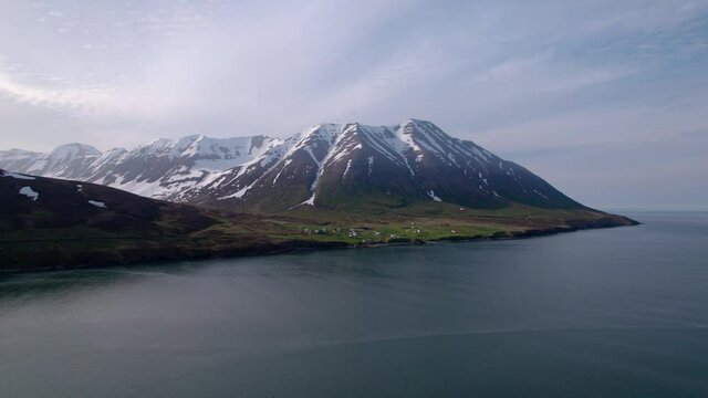 Epic aerial shot in Iceland of snowy mountains with small coastal village. Partly cloudy sky.