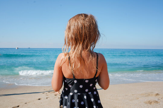 Toddler brown hair girl standing in front of the sea with her back to the camera. Beautiful sunny summer day.
