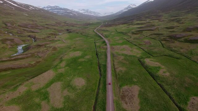 Aerial tracking shot of a red car driving through epic Icelandic landscape towards snowy mountains in the distance. Tracks perfectly with the car, on gravel road.