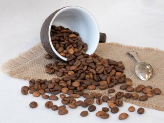 The coffee cup lies sideways on the table; grains of roasted aromatic coffee pour out of it onto a canvas napkin. Breakfast. Lunch break. Coffee business concept
