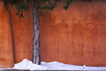 Obraz premium Tree trunk against an orange wall with snow piled on a sidewalk in Santa Fe, New Mexico 