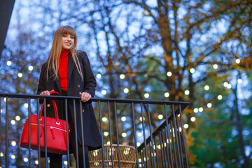 Happy young adult woman smiling with teeth smile, stays outdoors on city street at sunset time. Attractive girl weared in autumn clothes. Red bag, black coat. Blurred details