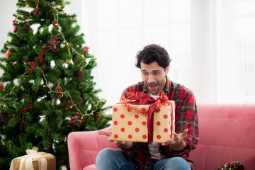 Obraz na płótnie Canvas happily handsome caucasian young man holding a gift box sitting on the sofa in a room decorated with Christmas trees and festive atmosphere in Christmas party.