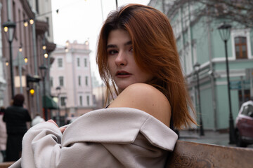 beautiful young girl in a beige coat sits on a bench in the city