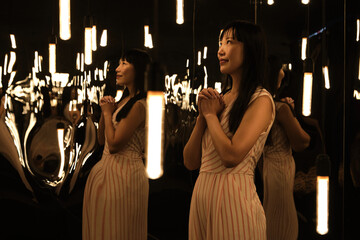 Young asian woman smiling and praying on a room full of lights and mirrors. © Ladanifer