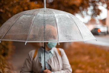 Old woman in bright clothes under a transparent umbrella and stretching hand catching water drops with content in bad weather in the city park