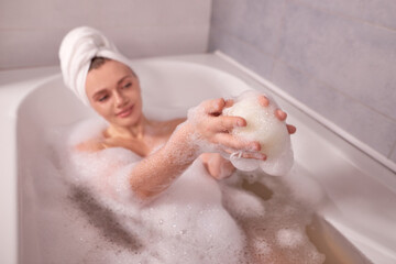 close up woman hands with shower sponge in soap foam bathtub at home. white towel in form of turban on head. shallow depth of field photo
