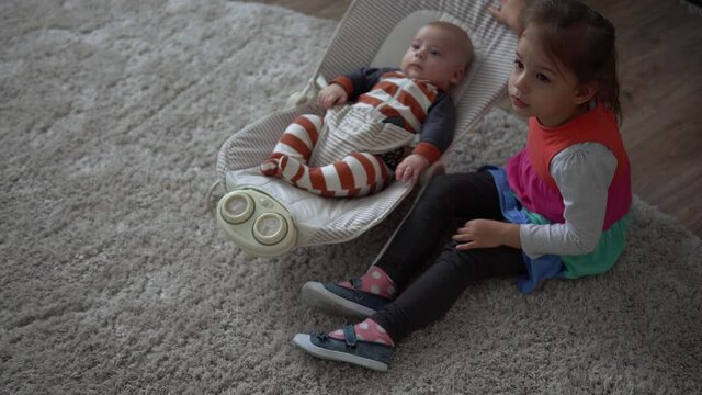 Childhood, motherhood, family, comfort. Little newborn baby boy in striped bodysuit pajamas awake looking around smiles plays rattles sibling sister kid lul amuse brother in room at home atmosphere