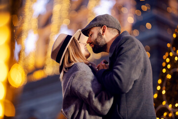 Fototapeta na wymiar Christmas couple kissing outdoors on New Year's eve. A young happy romantic couple standing on the street surrounded by Christmas lights, holding hands and kissing on New Year's eve.