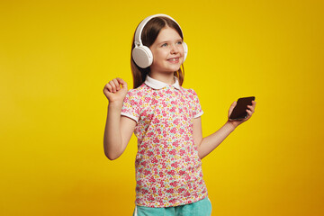 Adorable kid with white bluetooth headphones, listening music from smartphone and dancing, standing isolated over yellow background