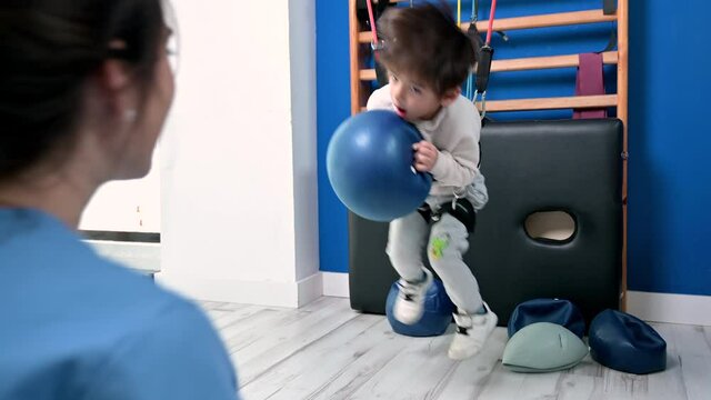 little kid with cerebral palsy has musculoskeletal therapy by doing exercises with a ball, in body fixing. Cheerful boy with disability at rehabilitation center for kids. High quality 4k footage