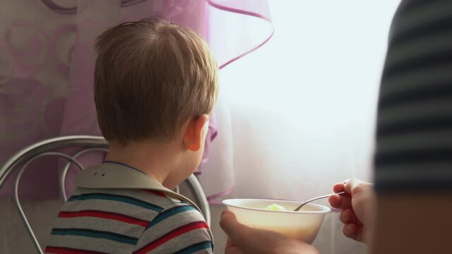 food, nutrition, childhood concepts - medium shot Mom feeding infant kid boy with rubber spoon. Cute hungry child dressed in bib sit in kids chair dont want to eats from plate indoors. dislike first