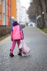 Little girl with a backpack, in a jacket and a hat near the school.