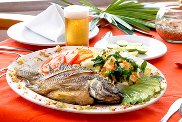 Mojarra, fried tilapia mojarra with lettuce, tomato and avocado salad accompanied by a glass of beer and lemons on a table in a Mexican restaurant