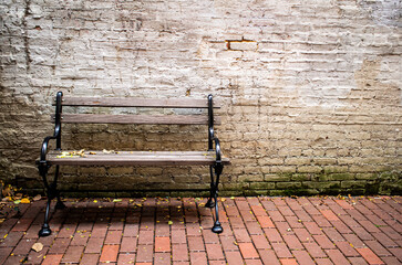 Weathered wood park bench in city park, dirty, crumbling white brick wall and red brick paving stones. Grainy, gritty, sad, aging feel. Autumn leaves scattered at the base of the wall. Copy Space.