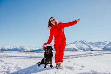 Attractive young brunette woman in red winter overalls and sun glasses posing with black Labrador retriever dog in the snowing mountains. Sunny day.