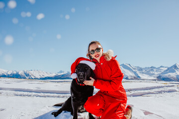 Attractive young brunette woman in red winter overalls and sun glasses posing with black Labrador retriever dog in the snowing mountains. Sunny day.