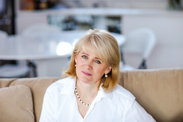 Portrait of beautiful adult blond woman in white shirt sitting on a sofa in the room. High quality photo