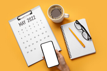 Female hand with mobile phone, clipboard with paper calendar for MAY 2022, cup of coffee, eyeglasses, pen and notebook on color background