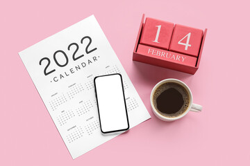Paper calendar for 2022 year and cube calendar with date FEBRUARY 14, mobile phone and cup of coffee on color background