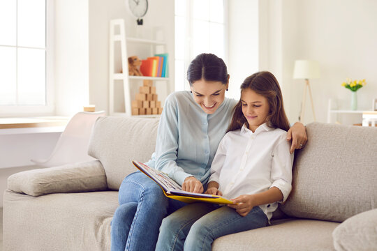 Joyful mother and her teenage daughter spend time together flipping through interesting book. Family sits on sofa at home in living room looking at family photo album or looking at pictures in book.