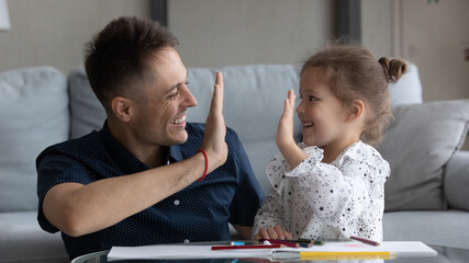 Happy excited dad and daughter girl giving high five over album with drawings, colorful pencils. Father showing praise, support for good homework result, playing creative learning games with child - obrazy, fototapety, plakaty