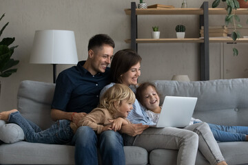 Happy joyful family watching online movie on laptop, relaxing on sofa together, enjoying internet TV channel, making video call. Millennial parents and two children resting at home