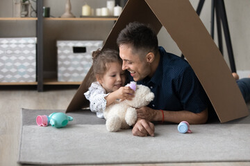 Happy playful daughter kid and loving daddy playing tea party in toy handmade house, relaxing on warm floor under cardboard paper roof, using childish colorful plastic dish. Childhood, housing