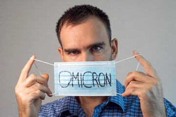 A man in a plaid shirt holds a medical mask with Omicron written on it in front of his face. Gray...