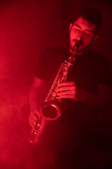 Fototapeta premium jeune musicien qui joue du saxophone. Ambiance cool jazz avec lumière rouge et fumée