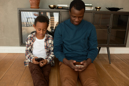 Communication gap. Dark-skinned father and his son sitting on floor in living-room and using smart phones, in silence, not noticing each other, having Internet and e-games addictions