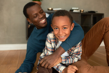 Fototapeta premium Human relationships. Positive emotions. Dark-skinned dad showing his love, pride and appreciation towards his son, hugging him sitting on floor, resting, laughing and enjoying bonding moment