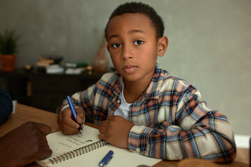 Portrait of adorable dark-skinned honor student doing homework, studying math, writing down...