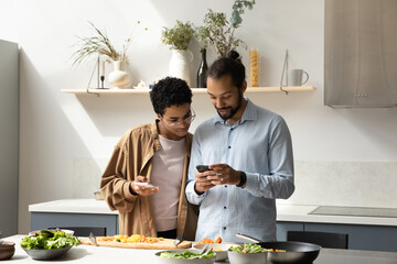 Distracted from cooking happy young African American family couple using cellphones, checking...