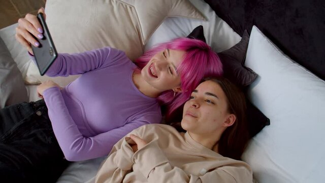 Top view of carefree joyful pink haired young woman and excited female friend with birth anomaly hands lying on bed, taking selfie shot on cellphone, making funny faces while relaxing at home.