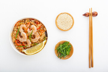 Rice with shrimps, vegetables and lemon in a paper disposable plate on a white table with chopsticks and wooden cups with sesame seeds and green onions. Asian street food. View from above