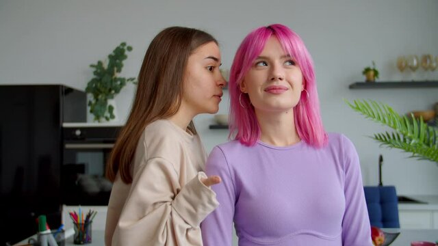 Portrait of cheerful lovely young woman with physical disability sharing a secret, whispering latest gossips to trendy amazed pink haired female friend while roommates relaxing in dorm room.
