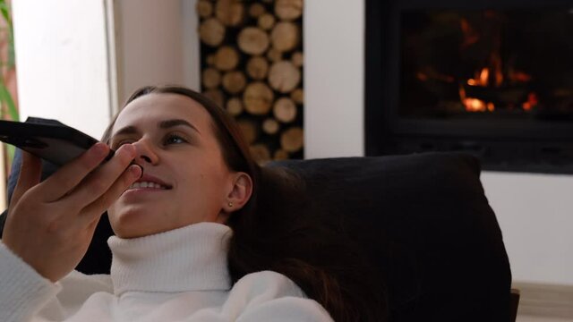 Relaxed young woman lying on sofa near fireplace, talking speaking dictating audio message in cellphone social network application, using ai voice virtual assistant web surfing information online