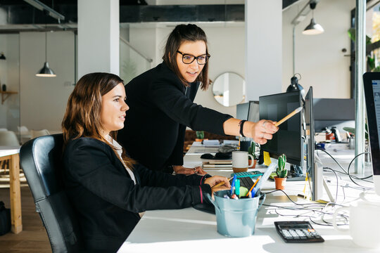 Smiling businesswoman listening to a collegue, in front of a computer, at desk, in office. They are in smart casuals.