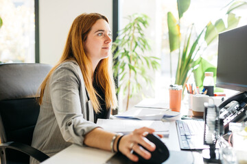 Young red haired woman, sitting on a desk and using a computer, in modern office