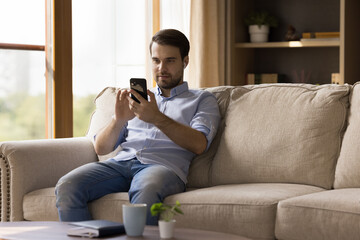 Obraz premium Millennial guy with earphones using media app on mobile phone at home, making video call, chatting online, watching webinar, listening to music, tunes from playlist. Man with earbud talking on cell