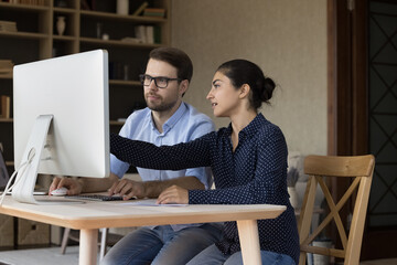 Two focused multiethnic coworkers working on project together, presentation on desktop monitor, sharing computer at workplace, discussing startup. Mentor training intern, explaining task